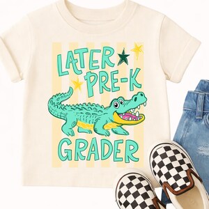 May include: A cream-coloured t-shirt with a cartoon alligator and the words "LATER PRE-K GRADER" in turquoise. The alligator is teal with a yellow belly and open mouth. The shirt is paired with denim jeans and checkered slip-on shoes.