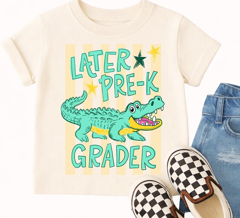 May include: A cream-coloured t-shirt with a cartoon alligator and the words "LATER PRE-K GRADER" in turquoise. The alligator is teal with a yellow belly and open mouth. The shirt is paired with denim jeans and checkered slip-on shoes.