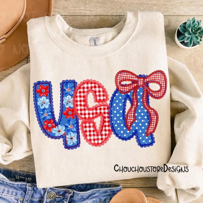 May include: Cream-colored sweatshirt with the letters "USA" in red, white, and blue. The "U" is blue with floral accents, the "S" is red gingham, and the "A" is blue with white polka dots. The text "ChouchoustoreDesigns" is printed on the bottom right.