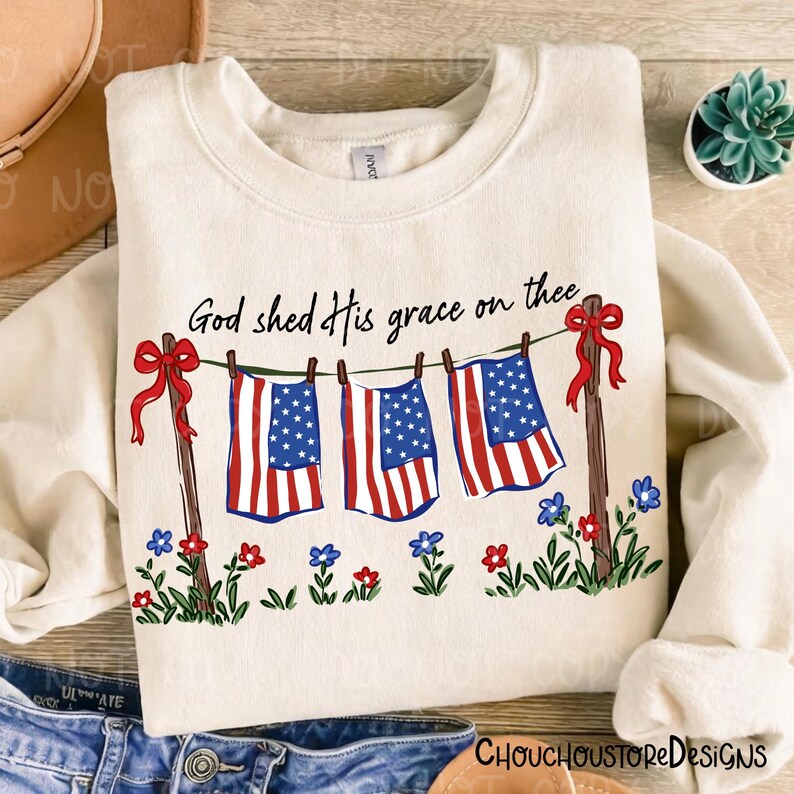 May include: Cream-colored sweatshirt with the text "God shed His grace on thee." The design features three American flags hanging on a clothesline, with red bows and floral accents. A patriotic theme.