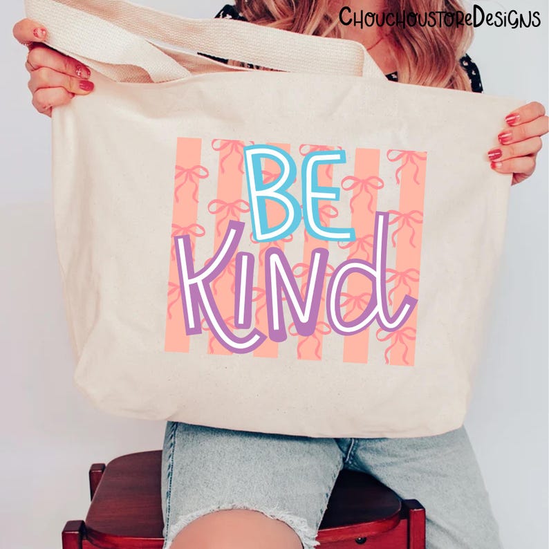 以下が含まれることがあります： 「BE KIND」の文字が青と紫で描かれたベージュのトートバッグ。背景には、ピンクのリボンがピーチ色のストライプに配置されています。バッグにはクリーム色のハンドルが付いています。