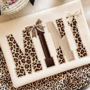 May include: Cream-colored fabric with the word "MINI" embroidered in a mix of leopard print, brown, and beige. The letters are outlined with lace and adorned with bows. The fabric is placed on a leopard print background.