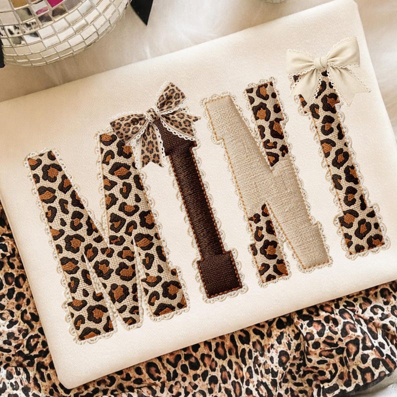 May include: Cream-colored fabric with the word "MINI" embroidered in a mix of leopard print, brown, and beige. The letters are outlined with lace and adorned with bows. The fabric is placed on a leopard print background.