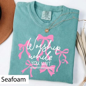 May include: Seafoam green t-shirt with the text "Worship while you wait" in white script and pink bow accents. A gold necklace with a pendant is draped over the shirt. The shirt is a Comfort Colors brand.