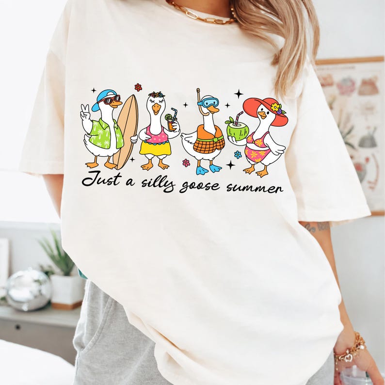 May include: Cream-colored t-shirt featuring a whimsical illustration of four cartoon geese in summer attire. One goose wears sunglasses and a hat, another holds a surfboard, and others have drinks. The text "Just a silly goose summer" is printed below.