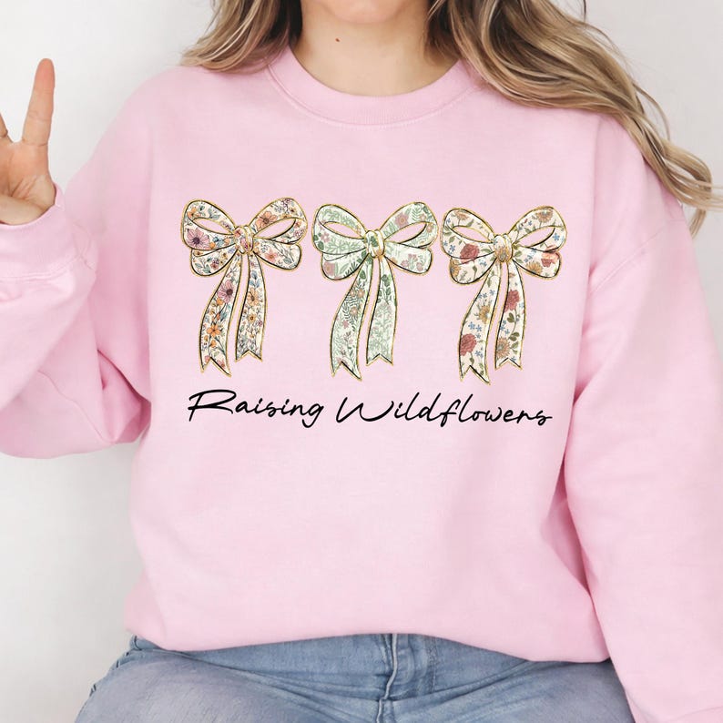 May include: Pink sweatshirt featuring three floral bows with gold outlines. The bows have a variety of floral patterns in shades of pink, green, and yellow. The text "Raising Wildflowers" is written in a cursive font below the bows.