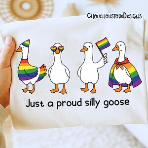 K&ouml;nnte beinhalten: Ein wei&szlig;es T-Shirt mit vier Cartoon-G&auml;nsen, die jeweils mit Regenbogen-Accessoires verziert sind. Die G&auml;nse tragen einen Regenbogenhut, herzf&ouml;rmige Sonnenbrillen, halten eine Regenbogenflagge und tragen einen Regenbogenumhang. Der Text lautet "Just a proud silly goose".