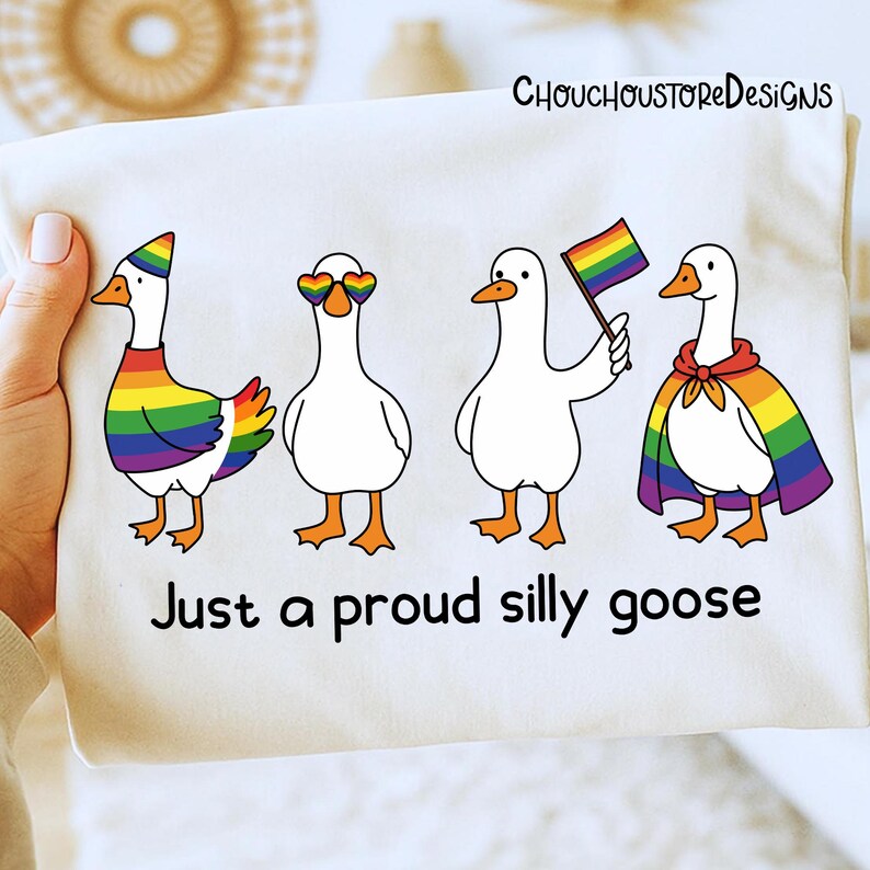 K&ouml;nnte beinhalten: Ein wei&szlig;es T-Shirt mit vier Cartoon-G&auml;nsen, die jeweils mit Regenbogen-Accessoires verziert sind. Die G&auml;nse tragen einen Regenbogenhut, herzf&ouml;rmige Sonnenbrillen, halten eine Regenbogenflagge und tragen einen Regenbogenumhang. Der Text lautet "Just a proud silly goose".
