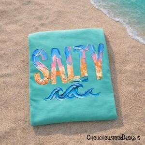 May include: A turquoise t-shirt with the word "SALTY" in colorful, textured lettering. Below the word are blue wave illustrations. The shirt is lying on a sandy beach near the ocean.