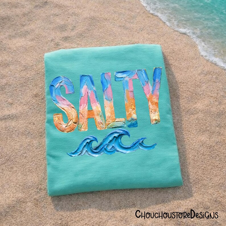 May include: A turquoise t-shirt with the word "SALTY" in colorful, textured lettering. Below the word are blue wave illustrations. The shirt is lying on a sandy beach near the ocean.