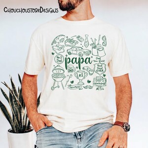 May include: Cream-colored t-shirt with a green doodle design. The word "papa" is in the center, surrounded by drawings of a car, grill, tie, and other dad-related items. The shirt has the text "No. 1 Dad", "Best Dad Ever", and "Dad's Workshop".