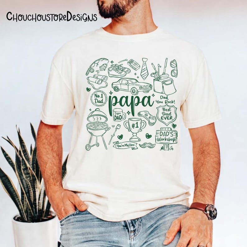 May include: Cream-colored t-shirt with a green doodle design. The word "papa" is in the center, surrounded by drawings of a car, grill, tie, and other dad-related items. The shirt has the text "No. 1 Dad", "Best Dad Ever", and "Dad's Workshop".