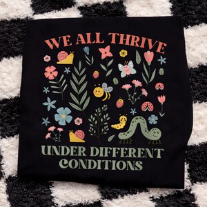 Peut inclure: T-shirt noir avec l'inscription "WE ALL THRIVE UNDER DIFFERENT CONDITIONS" en corail et vert. Le t-shirt pr&eacute;sente un motif floral et d'insectes color&eacute;s, notamment des fleurs, un papillon, une abeille, une coccinelle et une chenille.