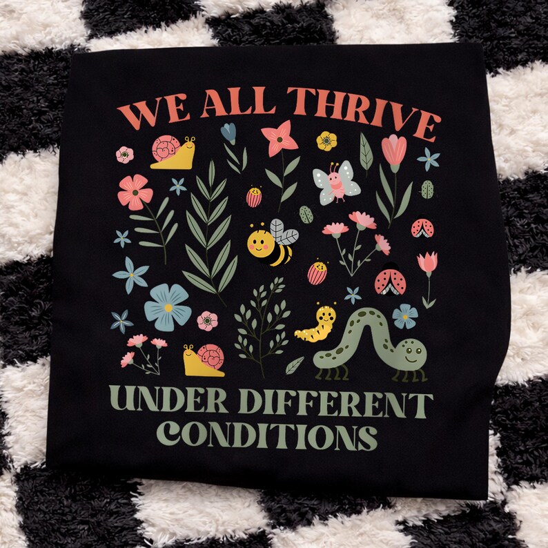 Peut inclure: T-shirt noir avec l'inscription "WE ALL THRIVE UNDER DIFFERENT CONDITIONS" en corail et vert. Le t-shirt pr&eacute;sente un motif floral et d'insectes color&eacute;s, notamment des fleurs, un papillon, une abeille, une coccinelle et une chenille.