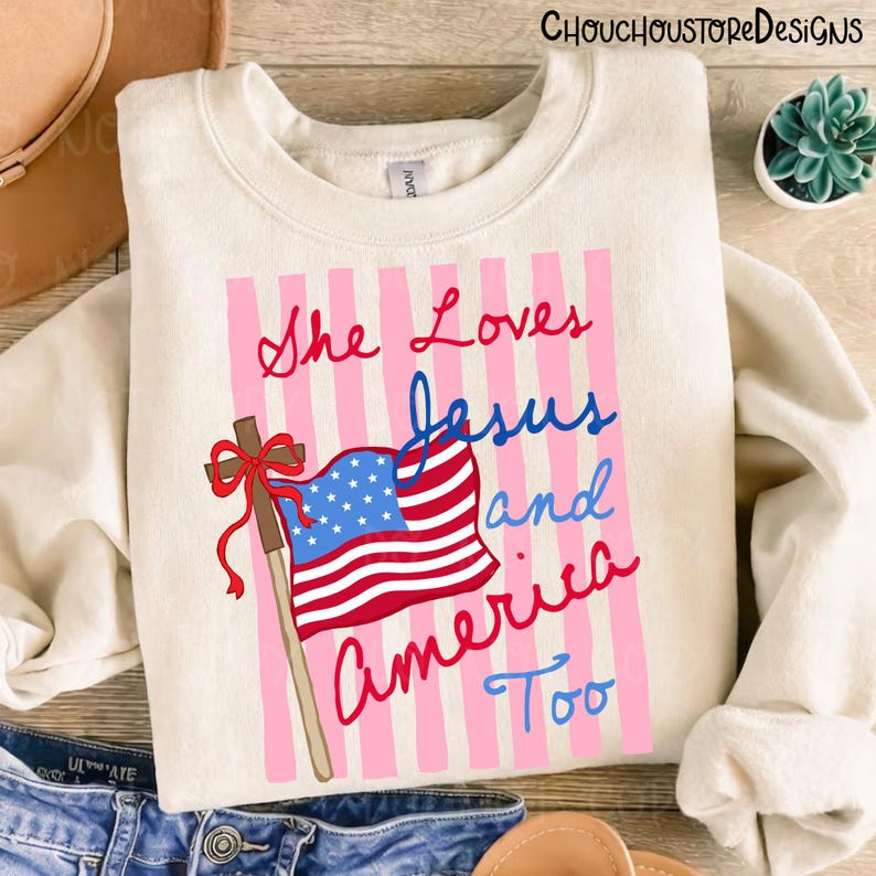 May include: Cream-colored sweatshirt with the text "She Loves Jesus and America Too" in red, blue, and pink. The design includes an American flag and a cross with a red bow, set against a pink striped background. A casual, patriotic theme.