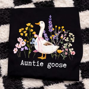 May include: Black t-shirt featuring a white goose with a pink bow, surrounded by colorful flowers. The text "Auntie goose" is printed in white below the illustration. The design is whimsical and nature-themed.
