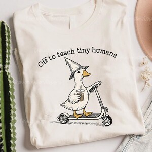 May include: A cream-colored t-shirt featuring a black ink illustration of a duck wearing a pointed hat, riding a scooter, and holding a coffee cup. The text above the duck reads, Off to teach tiny humans, in a simple black sans-serif font.