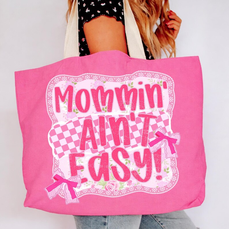 May include: A pink tote bag with the phrase "Mommin' Ain't Easy!" in a playful font. The design includes a checkered pattern, floral accents, and pink bows. The bag has cream-colored handles.