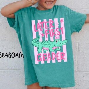May include: Teal t-shirt with a pink and white striped design. The shirt features the text "Later First Grader" and a cartoon alligator with a pink bow. The word "Seafoam" is printed on the left side of the shirt.