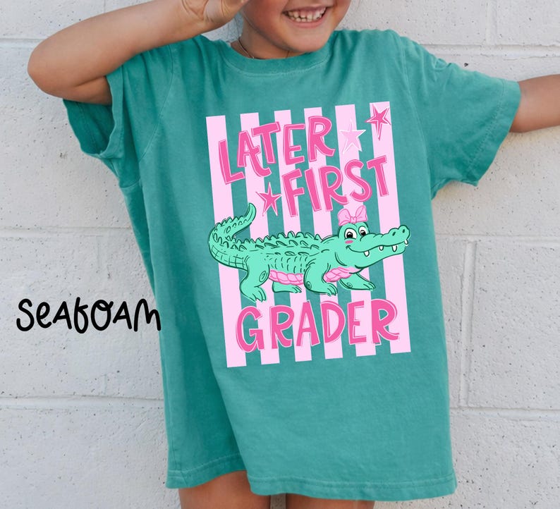 May include: Teal t-shirt with a pink and white striped design. The shirt features the text "Later First Grader" and a cartoon alligator with a pink bow. The word "Seafoam" is printed on the left side of the shirt.