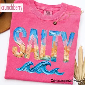 May include: A pink t-shirt with the word "SALTY" in a textured, colorful font, and blue wave graphics. The shirt has a crew neck and the brand "Comfort Colors" is visible. The text "crunchberry" is in the upper left corner.