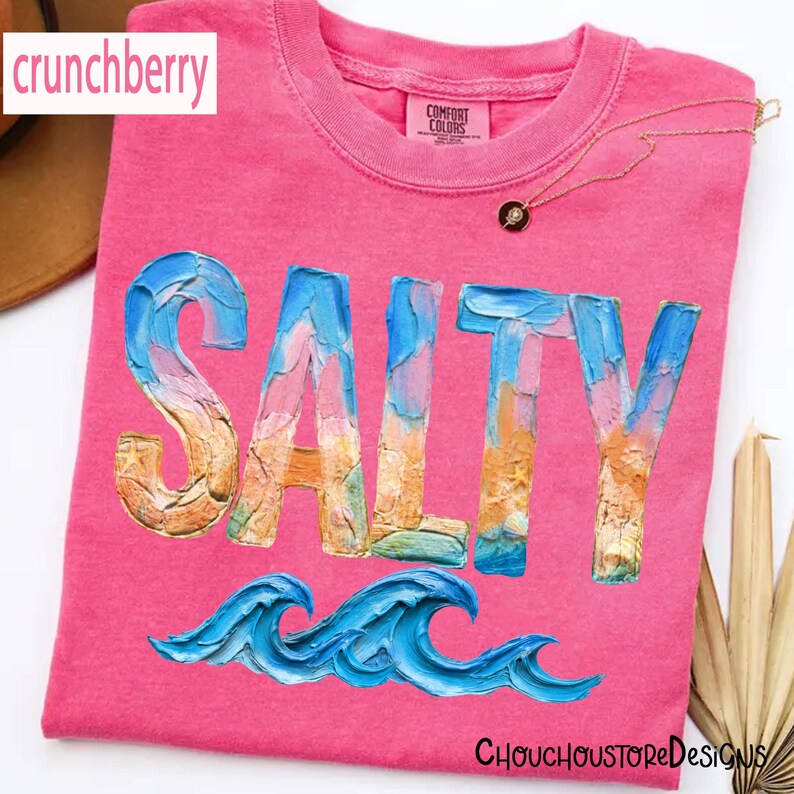 May include: A pink t-shirt with the word "SALTY" in a textured, colorful font, and blue wave graphics. The shirt has a crew neck and the brand "Comfort Colors" is visible. The text "crunchberry" is in the upper left corner.