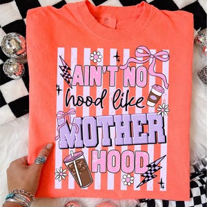 May include: Coral-colored t-shirt with the text "Ain't No Hood Like Motherhood" in a playful font. The design includes pink and purple accents, lightning bolts, bows, and coffee cup illustrations. A fun and colorful graphic tee.