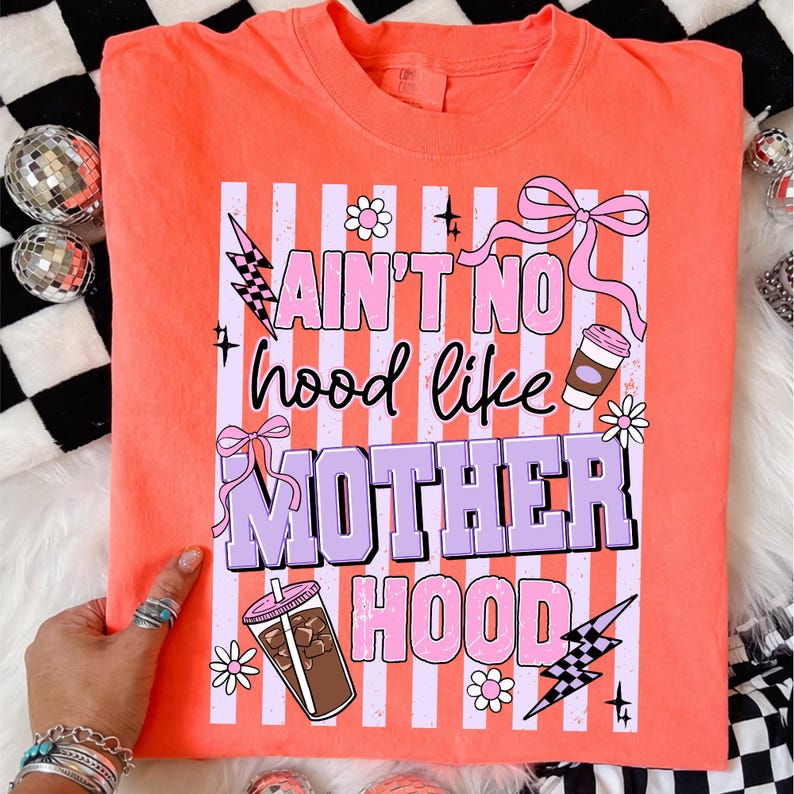 May include: Coral-colored t-shirt with the text "Ain't No Hood Like Motherhood" in a playful font. The design includes pink and purple accents, lightning bolts, bows, and coffee cup illustrations. A fun and colorful graphic tee.