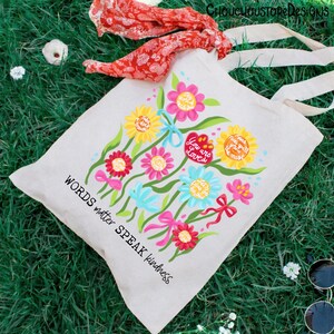 May include: A cream-colored tote bag with a floral design in various colors. The design includes flowers, bows, and the phrases "You are Loved" and "Words matter SPEAK kindness". The bag has long, cream-colored handles.