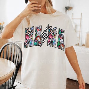 May include: Cream-colored t-shirt with the word "MAMA" in a colorful, decorative font. The letters are filled with floral and skull designs, and a black and white checkered lightning bolt is in the center.