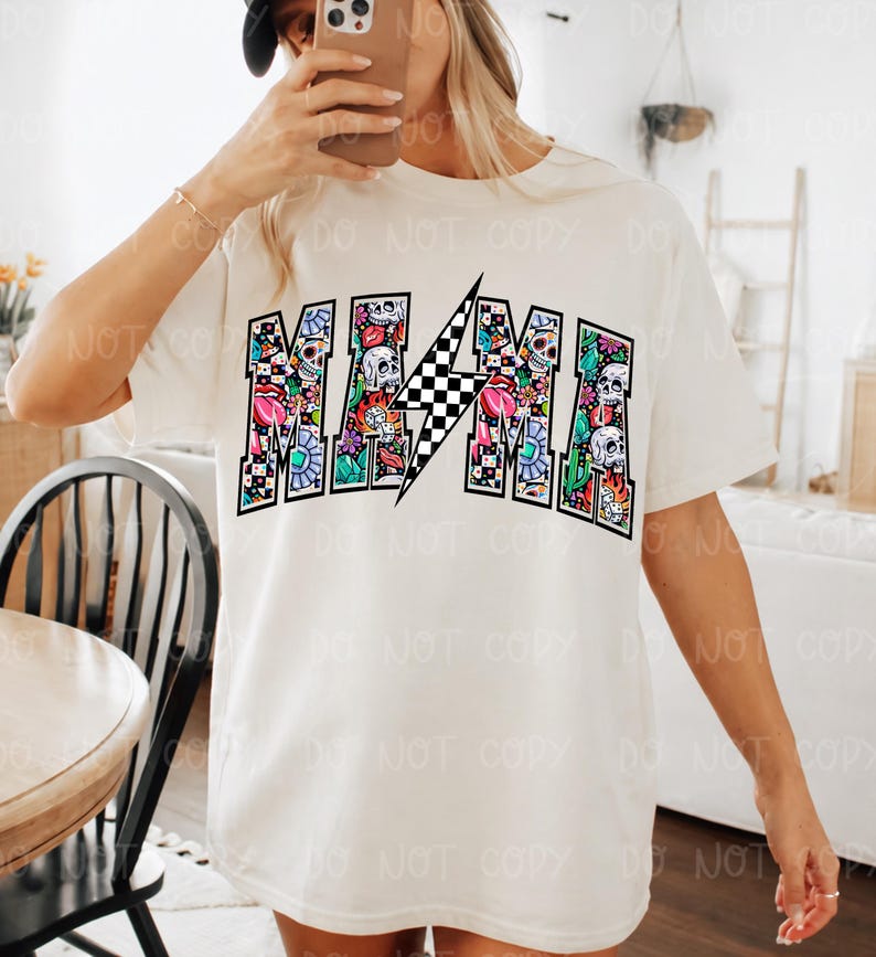 May include: Cream-colored t-shirt with the word "MAMA" in a colorful, decorative font. The letters are filled with floral and skull designs, and a black and white checkered lightning bolt is in the center.