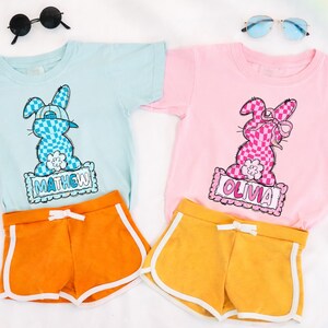 May include: Two children's t-shirts, one light blue and one pink, each with a cartoon bunny graphic. The light blue shirt has the name "MATHEW" and the pink shirt has the name "OLIVIA". Paired with orange and yellow shorts.