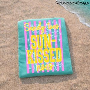 May include: A turquoise beach towel with pink vertical stripes and the words "Sandy toes, Sun-kissed noses" in yellow and white. A floral design is incorporated into the text. The towel is lying on a sandy beach.