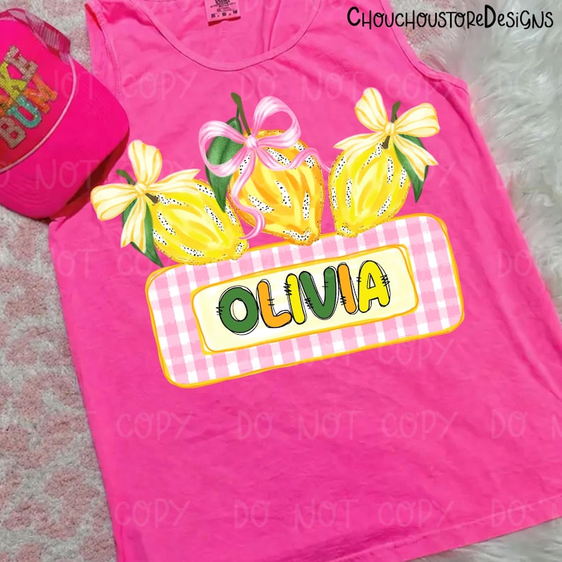 May include: A bright pink tank top featuring a lemon design with a pink and white gingham name tag that says "OLIVIA". The lemons have yellow and green details with pink bows. A pink baseball cap with the text "BUM" is visible.