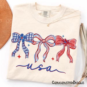 May include: Cream-colored t-shirt featuring three patriotic bows in red, white, and blue, with the word "usa" in blue script. The bows have different patterns, including gingham and polka dots. The shirt has a Comfort Colors tag.
