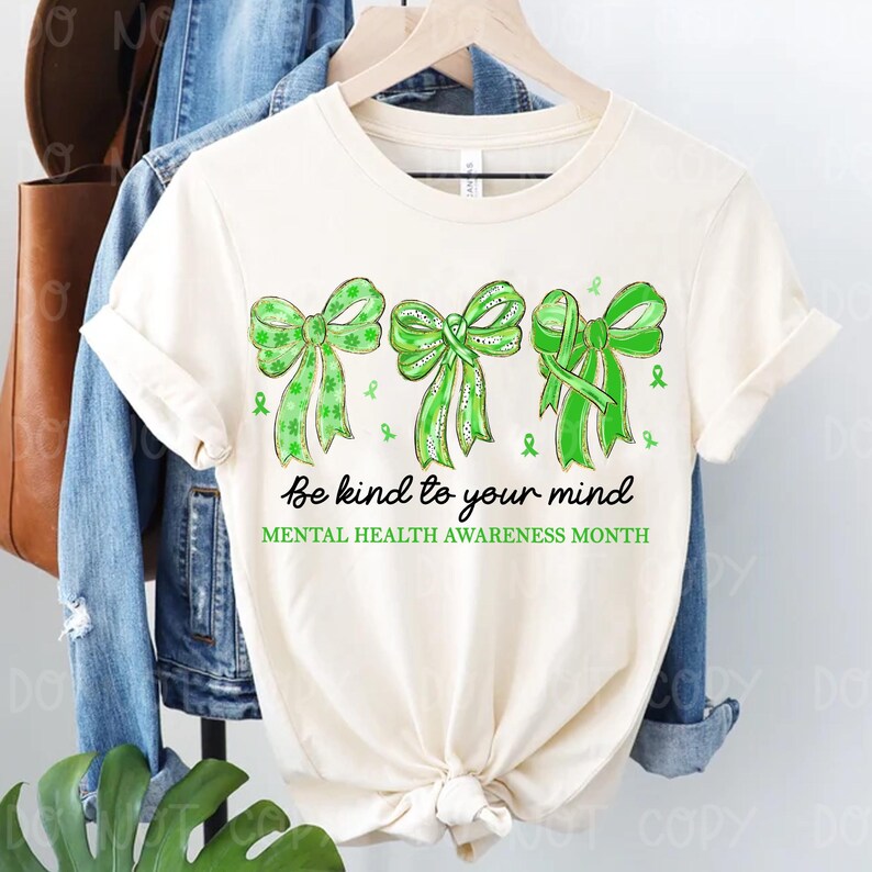 May include: Cream-colored t-shirt featuring three green bows and ribbons, with the text "Be kind to your mind" and "Mental Health Awareness Month". The bows have a watercolor effect. The shirt is hanging on a wooden hanger.