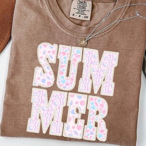 May include: A brown t-shirt with the word "SUMMER" in large, pastel-colored letters. The letters are filled with seashell and wave patterns. A silver necklace with a small pendant is on the shirt.
