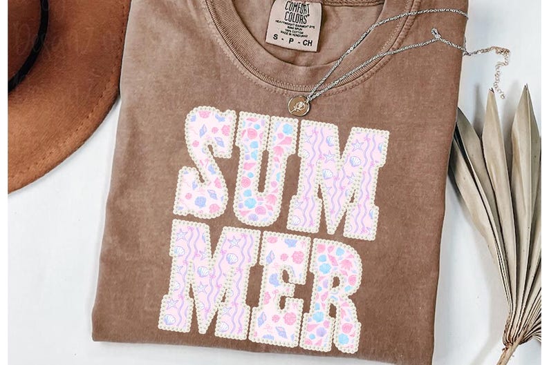 May include: A brown t-shirt with the word "SUMMER" in large, pastel-colored letters. The letters are filled with seashell and wave patterns. A silver necklace with a small pendant is on the shirt.