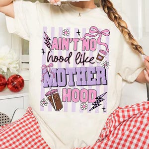 May include: Cream-colored t-shirt with a graphic design. The design features the text "Ain't No Hood Like Motherhood" in pink and purple, along with illustrations of coffee cups, flowers, and lightning bolts. The shirt has a relaxed fit.