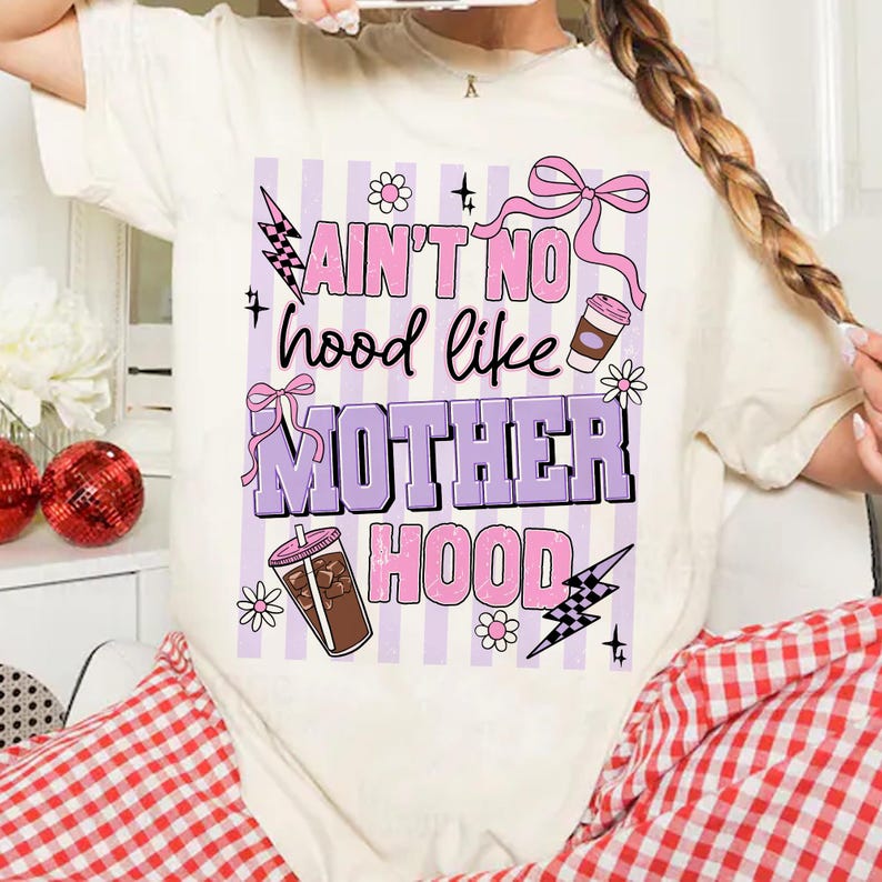 May include: Cream-colored t-shirt with a graphic design. The design features the text "Ain't No Hood Like Motherhood" in pink and purple, along with illustrations of coffee cups, flowers, and lightning bolts. The shirt has a relaxed fit.