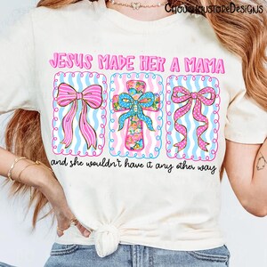 May include: Cream-colored t-shirt with the text "JESUS MADE HER A MAMA" in pink. The design includes three framed images: two pink bows and a floral cross, all set against a blue and white striped background. The phrase "and she wouldn't have it any other way" is below.