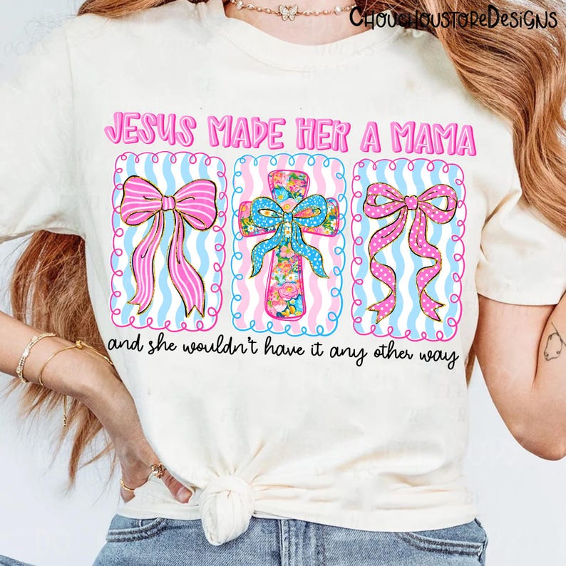 May include: Cream-colored t-shirt with the text "JESUS MADE HER A MAMA" in pink. The design includes three framed images: two pink bows and a floral cross, all set against a blue and white striped background. The phrase "and she wouldn't have it any other way" is below.