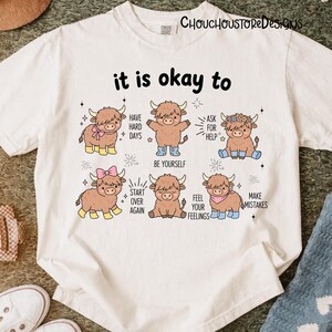 May include: White t-shirt with the text "it is okay to" above illustrations of cartoon Highland cows. Each cow has a different accessory and message, including "Have Hard Days" and "Be Yourself."
