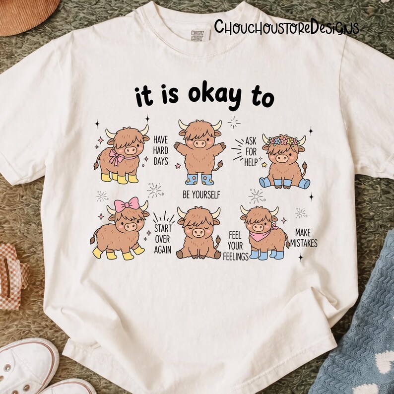 May include: White t-shirt with the text "it is okay to" above illustrations of cartoon Highland cows. Each cow has a different accessory and message, including "Have Hard Days" and "Be Yourself."