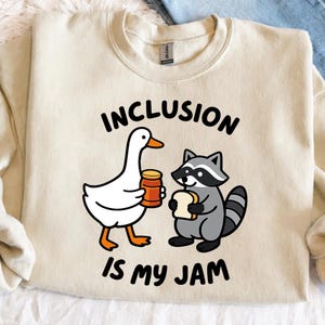 May include: Cream-colored sweatshirt with the text "INCLUSION IS MY JAM." The graphic features a cartoon goose holding a jar and a raccoon holding bread. The goose is white with orange feet and beak, and the raccoon is gray with a striped tail.