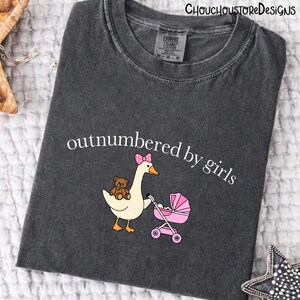 May include: Dark gray t-shirt with the text "outnumbered by girls." The graphic features a cartoon goose with a pink bow, a teddy bear, and a pink stroller. The shirt is made of a soft, comfortable material.