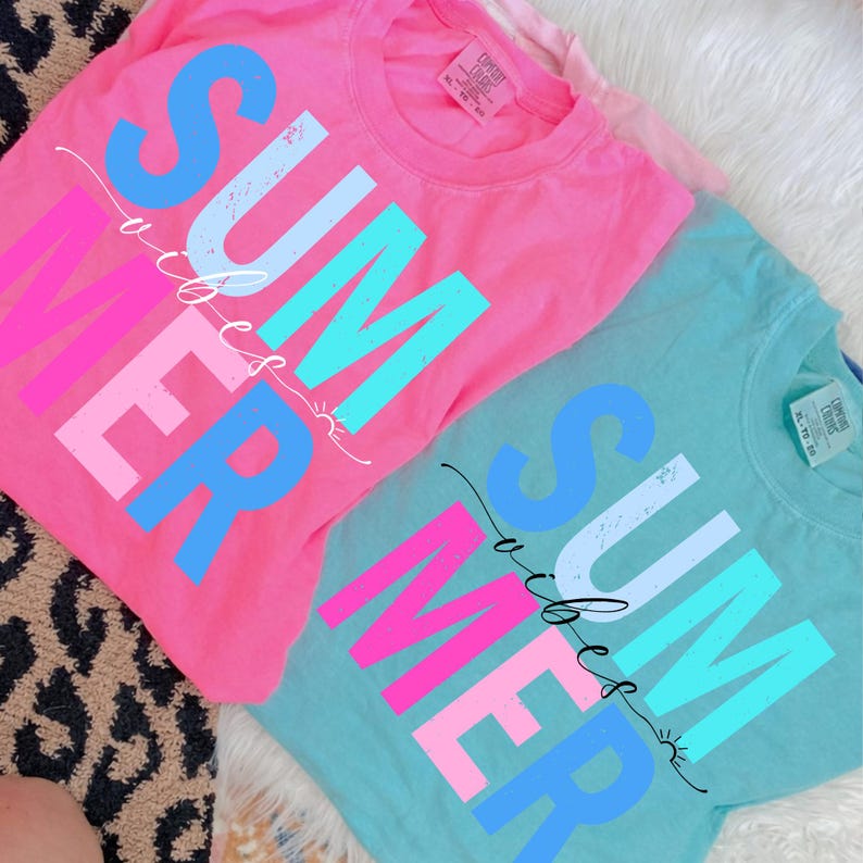May include: Two t-shirts, one pink and one teal, with the word "SUMMER" in large, colorful letters. The word "SUMMER" is in a mix of blue, pink, and white. A cursive line with the word "vibes" is written across the word "SUMMER".