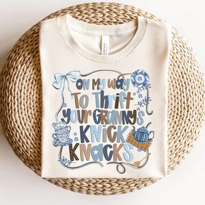 May include: Cream-colored t-shirt with the text "On My Way To Thrift Your Granny's Knick Knacks" in blue and brown lettering. The design includes illustrations of a bow, teacups, and a teapot.