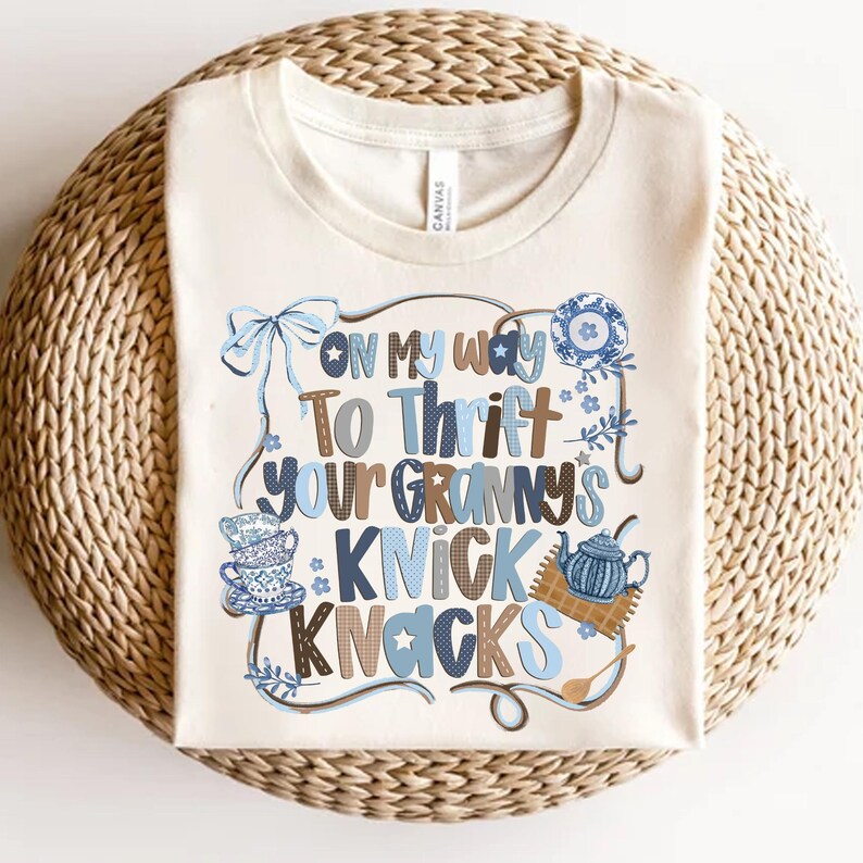 May include: Cream-colored t-shirt with the text "On My Way To Thrift Your Granny's Knick Knacks" in blue and brown lettering. The design includes illustrations of a bow, teacups, and a teapot.