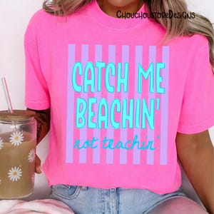 May include: A bright pink t-shirt with the text "CATCH ME BEACHIN' not teachin'" in turquoise and purple. The shirt has vertical purple stripes behind the text. The shirt is a casual wear item.